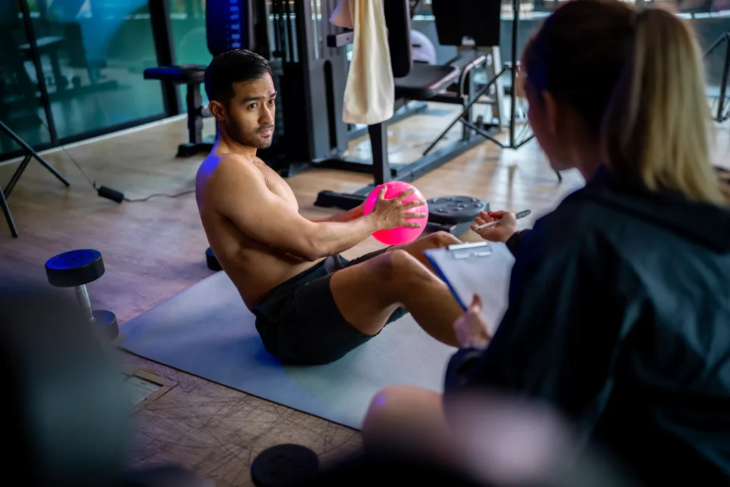 Fitness trainer guiding a male client performing sit-ups with a ball in the gym while taking notes.
