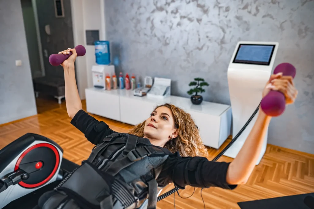 Woman performing a strength training exercise with dumbbells while wearing an EMS (electrical muscle stimulation) suit in a gym.