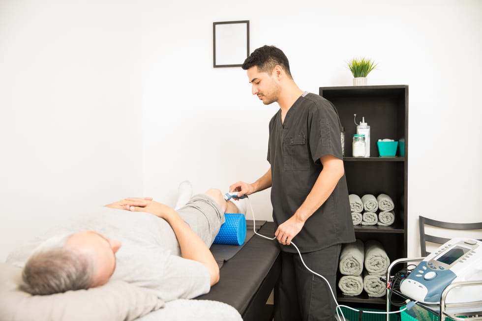 Physical therapist using an ultrasound probe on a patient's knee while standing in a hospital.